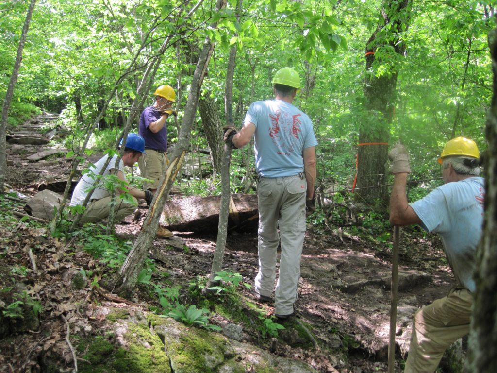 Four men in hard hats move a large stone on the trail using  a winch