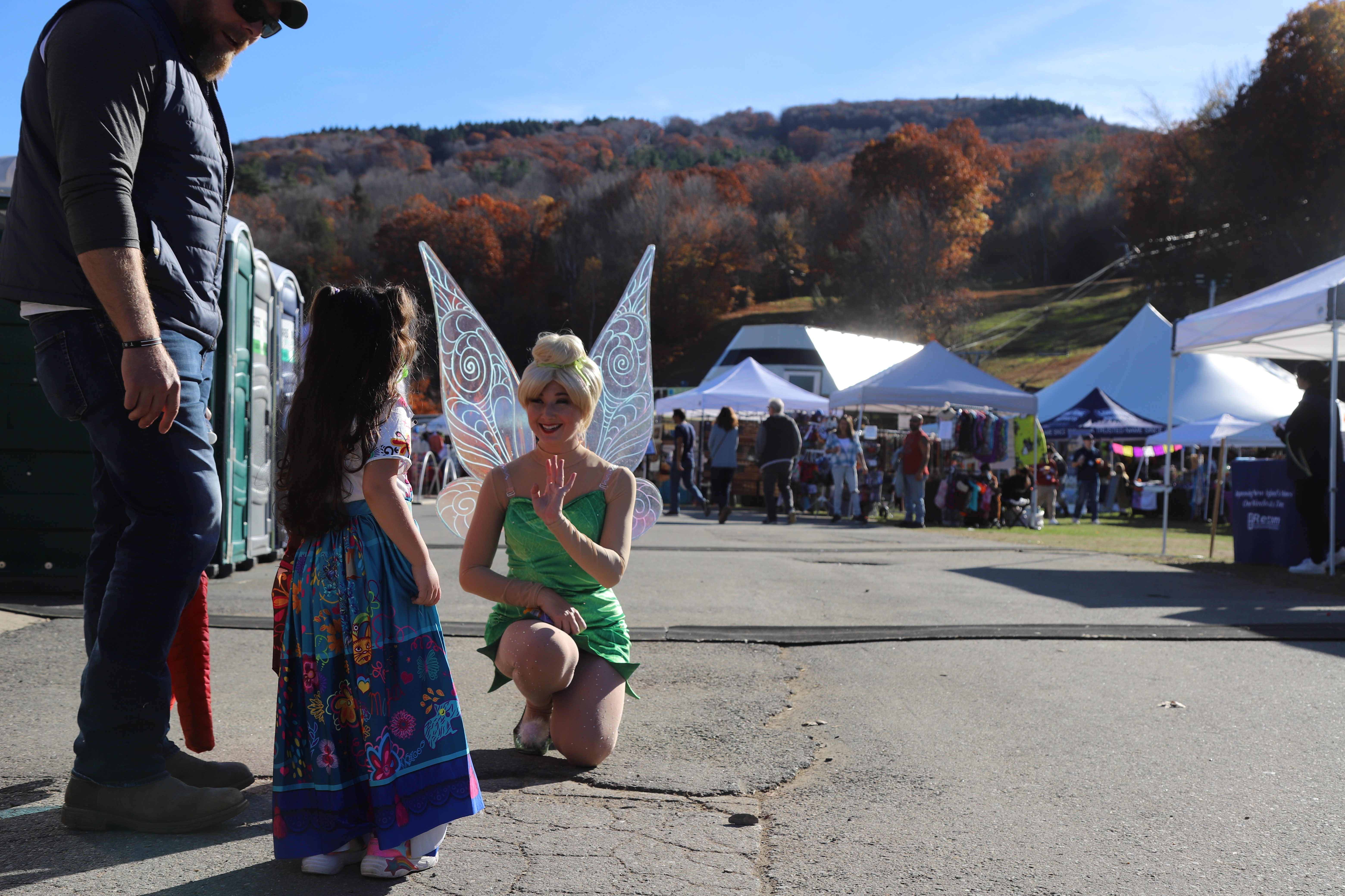 A woman dressed as Tinkerbell kneels to greet a little girl on the front road of an outdoor festival